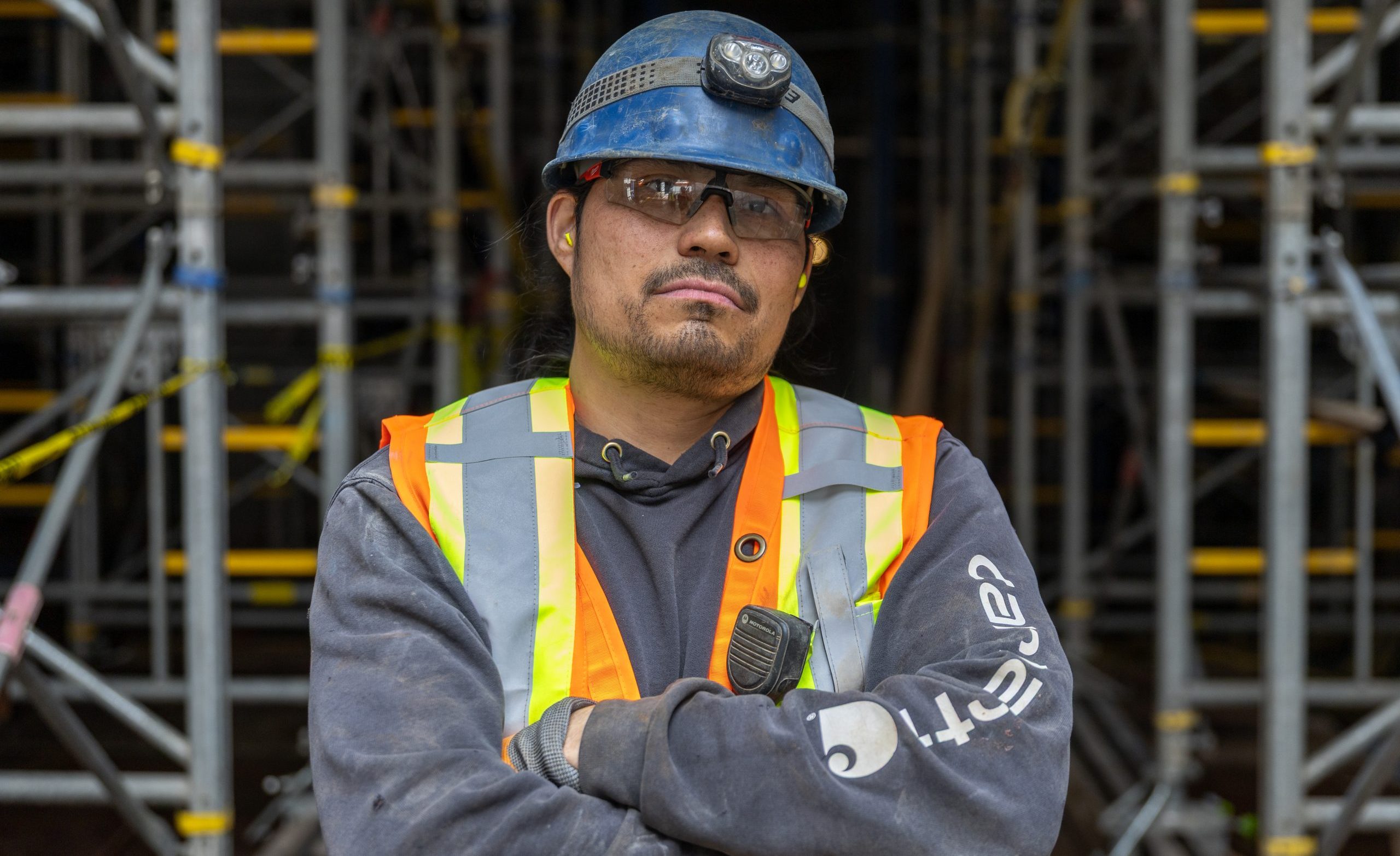 Picture off a construction worker looking at the camera, hands crossed, with scaffolding behind him.